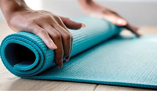 Mains qui roulent un tapis de yoga turquoise sur un plancher de bois clair